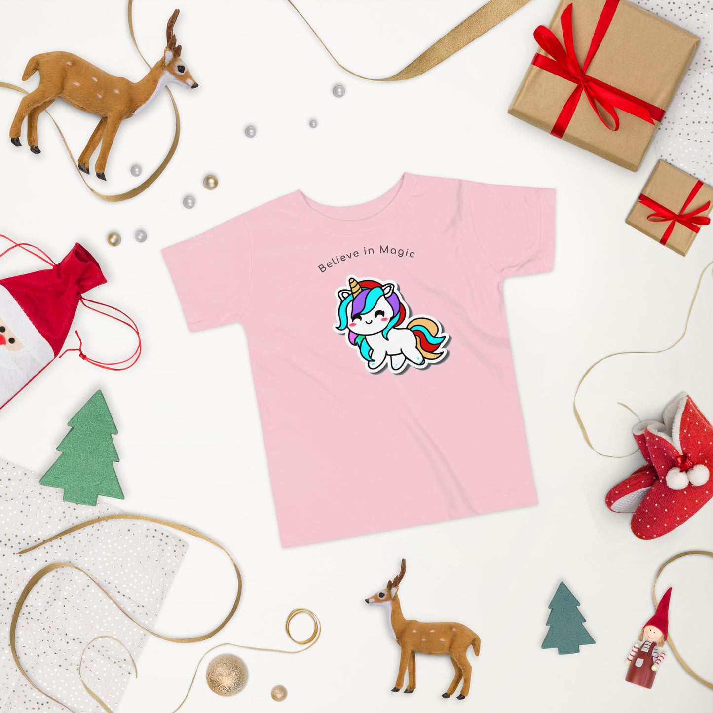 pink t-shirt with smiling and colorful unicorn laid on a white surface surrounded by Christmas items
